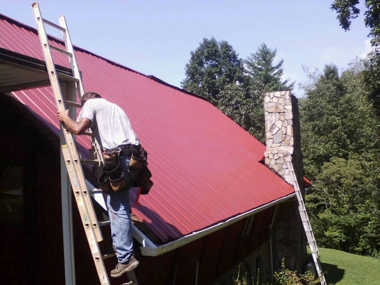 Roofer on a ladder beside a red metal roof and stone chimney — Soffit & Fascia Repair by Roofing Brothers in Belle Chasse